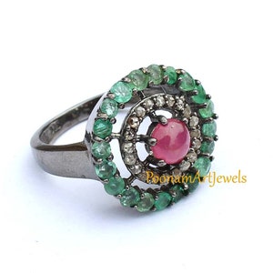 May include: A sterling silver ring featuring a central round ruby gemstone, surrounded by a halo of small diamonds and a second halo of emeralds. The ring has a dark, oxidized finish and is set against a white background.