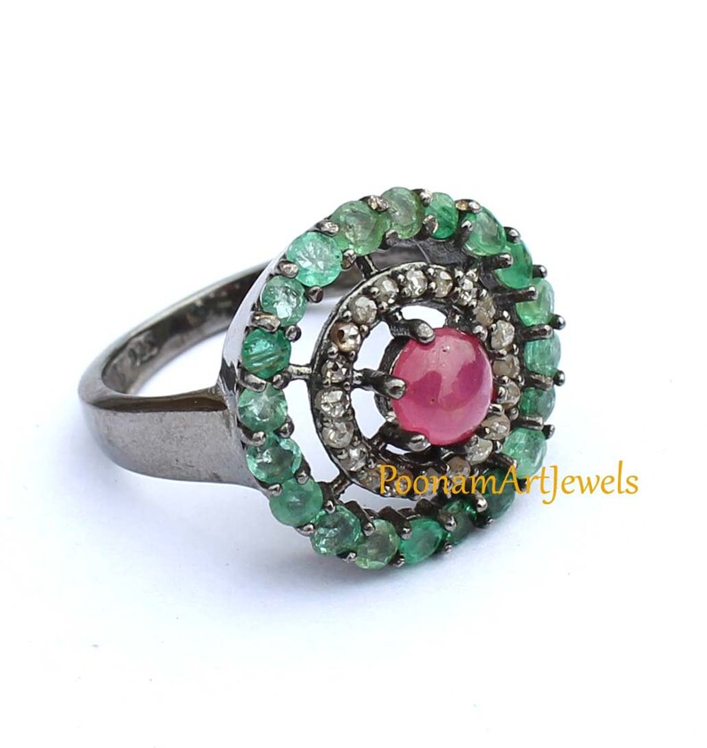 May include: A sterling silver ring featuring a central round ruby gemstone, surrounded by a halo of small diamonds and a second halo of emeralds. The ring has a dark, oxidized finish and is set against a white background.