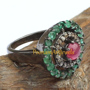 May include: A close-up of a ring with a dark metal band. The ring features a central red gemstone, surrounded by smaller clear and green gemstones. The ring is set on a wooden surface. The text "PoonamArtJewels" is visible.