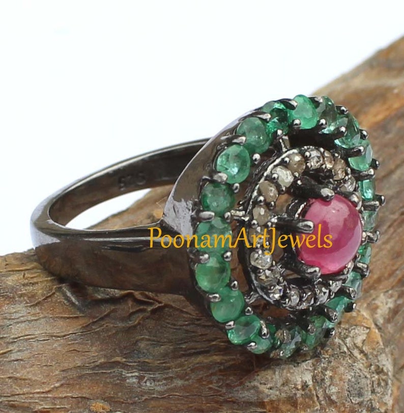 May include: A close-up of a ring with a dark metal band. The ring features a central red gemstone, surrounded by smaller clear and green gemstones. The ring is set on a wooden surface. The text "PoonamArtJewels" is visible.
