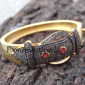 Puede incluir: Pulsera de oro con un diseño de hebilla engastada con diamantes negros y blancos. La hebilla tiene dos pequeñas piedras preciosas rojas. La pulsera está etiquetada como "Poonam Artjewels".