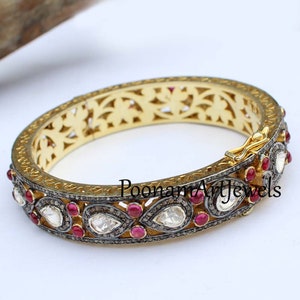 Könnte beinhalten: Goldarmband mit filigranem Design und besetzt mit birnenförmigen Diamanten und roten Rubinen. Das Armband hat einen Scharnierverschluss und ist mit "Poonam Art Jewels" signiert.