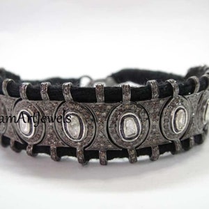 May include: A black cord bracelet with silver accents and round, white gemstones. The bracelet is set with small diamonds and features the text "Poonam Art Jewels".