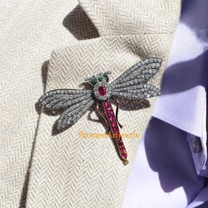 May include: A silver dragonfly brooch with ruby and emerald accents. The dragonfly is set with sparkling diamonds and has a long, slender body.