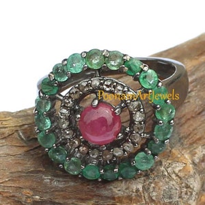 May include: A silver ring featuring a central, round, red gemstone surrounded by a halo of small, clear stones and a ring of green gemstones. The ring is set against a natural wood background. The text "PoonamArtJewels" is visible.