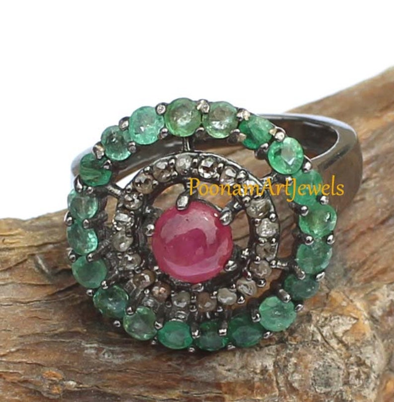 May include: A silver ring featuring a central, round, red gemstone surrounded by a halo of small, clear stones and a ring of green gemstones. The ring is set against a natural wood background. The text "PoonamArtJewels" is visible.