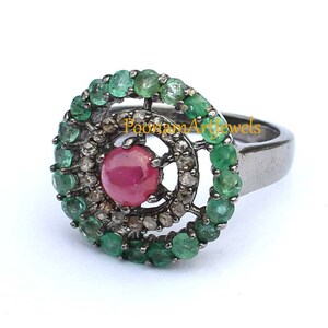 May include: A silver ring featuring a central ruby gemstone surrounded by a halo of small diamonds and a border of emeralds. The ring has a dark, oxidized finish and a classic design.