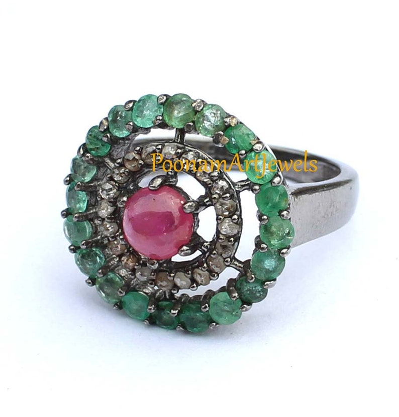 May include: A silver ring featuring a central ruby gemstone surrounded by a halo of small diamonds and a border of emeralds. The ring has a dark, oxidized finish and a classic design.