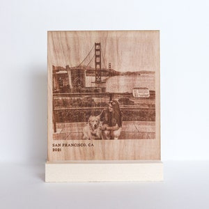 May include: A wooden plaque with a photo of the Golden Gate Bridge in San Francisco, California. The photo shows a woman and a dog sitting on a bench in front of the bridge. The plaque is engraved with the text "San Francisco, CA 2021".
