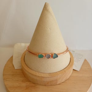 May include: A rose gold bangle bracelet featuring three small, raw, irregularly shaped stones in shades of green, light purple, and blue, set horizontally along the centre of the thin, polished metal band.