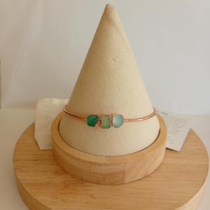 May include: A thin copper-toned metal bangle bracelet featuring three small, smooth, frosted sea glass pieces in varying shades of green. The bracelet is displayed on a beige cone-shaped stand, showcasing the vibrant green glass elements centred on the wire.