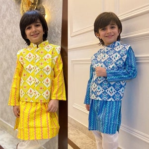 May include: Two young boys wearing traditional Indian clothing. The boy on the left is wearing a yellow kurta with a white and yellow patterned vest. The boy on the right is wearing a blue kurta with a white and blue patterned vest. Both boys are wearing white pants and brown shoes.