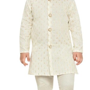 May include: A young boy wearing a white kurta with gold embroidery and matching white trousers. He is wearing gold shoes.
