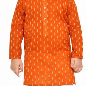 May include: A young boy wearing a long-sleeved orange kurta with a white pattern and white trousers. He is wearing gold shoes.