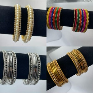 May include: Set of four bangle bracelets. The first set is silver with clear rhinestones. The second set is gold with clear rhinestones. The third set is white with pearls. The fourth set is multi-colored thread with gold accents.
