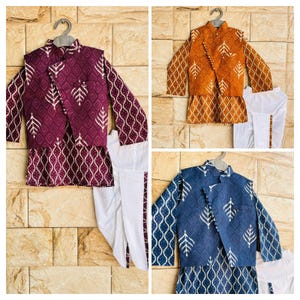 Può includere: Tre completi di abbigliamento etnico per bambini, ciascuno con una giacca fantasia, una camicia a maniche lunghe e pantaloni bianchi. Le giacche sono bordeaux, arancioni e blu, con motivi a foglie e diamanti. I pantaloni hanno una striscia decorativa.