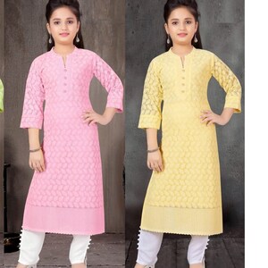 May include: Two girls wearing matching outfits. The girl on the left is wearing a pink long-sleeved kurta with white pants and the girl on the right is wearing a yellow long-sleeved kurta with white pants. Both girls are wearing white shoes.