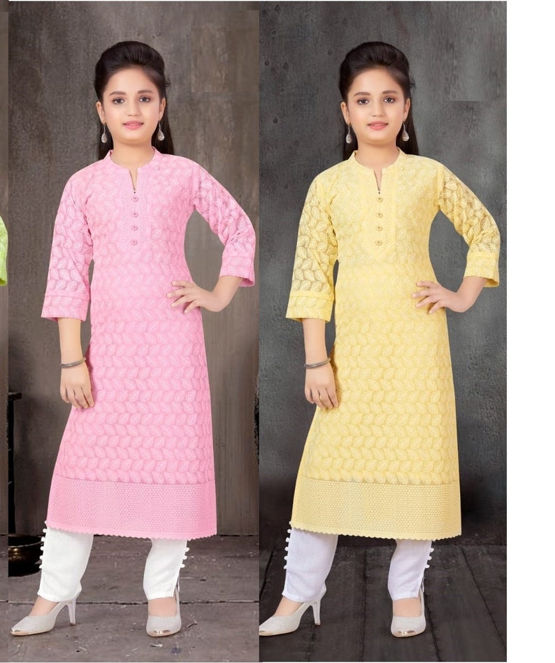 May include: Two girls wearing matching outfits. The girl on the left is wearing a pink long-sleeved kurta with white pants and the girl on the right is wearing a yellow long-sleeved kurta with white pants. Both girls are wearing white shoes.