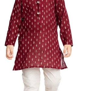May include: A young boy wearing a maroon kurta with a white print and white trousers. He is wearing white shoes.