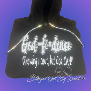 May include: Black hoodie with the text "God-fi-dence" and "Knowing I can't, but God CAN!" in white lettering. The text is outlined with rhinestones. The hoodie also has the text "be blessed" in white lettering with a rhinestone infinity symbol. The text "Blinged Out By Bella" is also included in white lettering with a rhinestone star.