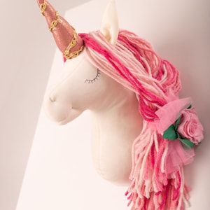 May include: A plush unicorn head with a pink and gold horn, pink yarn mane, and a pink rose accent. The unicorn is white with closed eyes and is mounted on a white surface. The horn is adorned with gold trim.