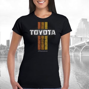 May include: Black t-shirt with a vintage-style graphic of the word "TOYOTA" in white with a distressed look. The graphic is surrounded by four vertical orange and yellow stripes. The text "4RUNNER" is printed in white below the graphic.