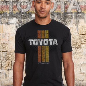 May include: A black t-shirt with a vintage-style graphic of the word "TOYOTA" in white with a gray distressed effect. The graphic also includes three vertical stripes in orange, yellow, and brown, and the text "4RUNNER" in white below the word "TOYOTA".
