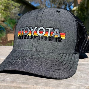 Puede incluir: Una gorra de camionero negra y gris con una parte trasera de malla negra. La gorra tiene un logotipo bordado en blanco que dice "TOYOTA" con una franja arcoíris debajo.