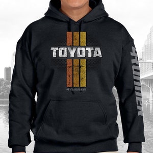 May include: Black hooded sweatshirt with a distressed graphic design featuring the word "TOYOTA" in white with orange and yellow stripes and the text "4RUNNER" in white below. The left sleeve has the word "4RUNNER" printed vertically in white.