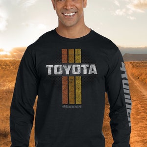 May include: Black long-sleeve t-shirt with a vintage-style graphic of the word "TOYOTA" in white with a distressed look. The graphic also includes three vertical stripes in orange, brown, and yellow, with the words "4RUNNER" in white below the stripes.