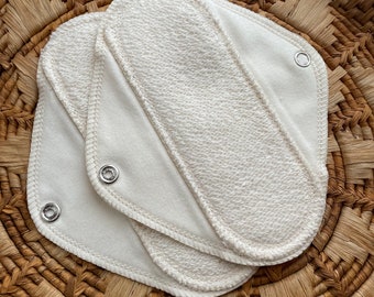 May include: Three off-white reusable cloth pads featuring a textured terry cloth center panel and smooth fabric wings. Each pad is secured with a silver-toned metal snap fastener at the corners, showcasing a layered design with stitched edges.
