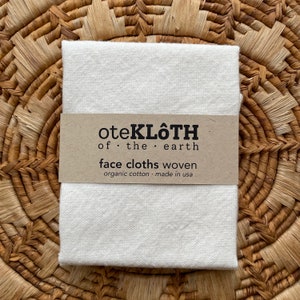 以下が含まれることがあります： 茶色のラベルに「oteKLÖTH of the earth face cloths woven organic cotton made in USA」と書いてある、白いオーガニックコットン製のフェイスクロスです。