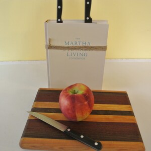 May include: A wooden cutting board with a red apple and a silver knife. The cutting board has alternating stripes of light and dark wood. The book "The Martha Stewart Living Cookbook" is in the background.