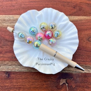May include: A cream-colored pen with a silver tip and the text "The Crafty PersimmonPig" is displayed on a white, ruffled dish. The dish holds several colorful, iridescent beads with cartoon animal designs.