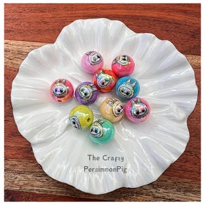 May include: A collection of colorful, round beads with cartoon bunny faces, arranged on a white, scalloped dish. The beads are in various colors, including pink, blue, and yellow. The dish has the text "The Crafty PersimmonPig".