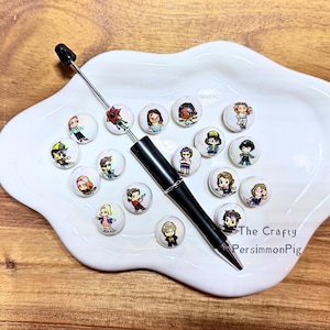May include: A collection of iridescent cabochons featuring cartoon characters, arranged on a white, wavy-edged dish. A black pen with a silver tip rests among the cabochons. The text "The Crafty PersimmonPig" is visible on the dish.