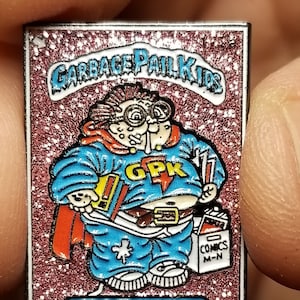 May include: A Garbage Pail Kids pin featuring Nat Nerd, a cartoon character with a red cape, blue jeans, and a blue shirt with the letters GPK. The pin has a pink glitter background and the text "Garbage Pail Kids" and "Nat Nerd".