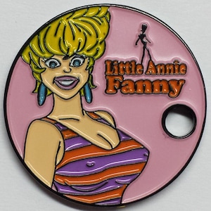 May include: A round, pink enamel pin with a black outline. The pin features a cartoon woman with blonde hair wearing a striped swimsuit. The text "Little Annie Fanny" is written in orange letters on the pin.