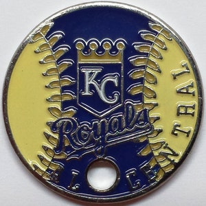May include: A silver baseball-themed coin with a blue and yellow enamel design. The coin features the Kansas City Royals logo, including the team's name and a crown. The coin has a hole in the center.