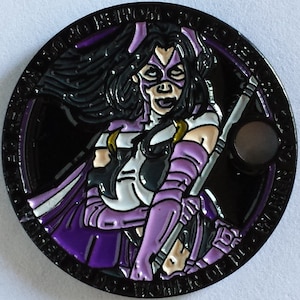 May include: A black and purple metal coin featuring a comic book character with a purple mask and a staff. The coin has a black background with a circle in the center. The text around the edge of the coin reads "The Huntress is Coming".