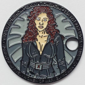 May include: A black and silver metal coin featuring a comic book character with red hair and a black outfit. The character is standing in front of a shield with a star in the center. The coin has the text "Marvel Comics" around the edge.