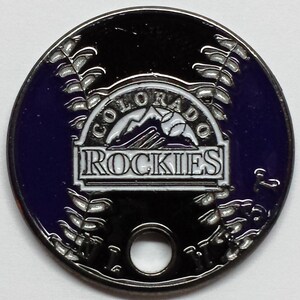 May include: A silver and purple metal coin with a white logo of the Colorado Rockies baseball team. The logo features a mountain range and the words "Colorado Rockies".