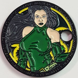 May include: A black and green metal coin with a woman's face in the center. The woman has long black hair and is wearing a green outfit. The coin has a yellow and black design around the edge.