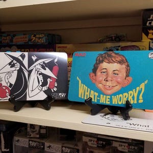 Puede incluir: Dos letreros decorativos en exhibición. Uno presenta a los personajes de Spy vs. Spy con una bomba, el otro a Alfred E. Neuman con la frase "What, me worry?" sobre un fondo azul. Los letreros están en un estante.