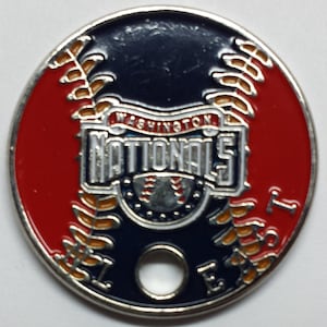 May include: A silver metal baseball coin with a red and blue design. The coin features the Washington Nationals logo with the team name and a baseball. The coin has a hole in the center for hanging.