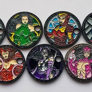 May include: Set of eight round metal tokens featuring different female superhero characters. Each token has a black border and a colorful image of a superhero with a unique design. The tokens have a hole in the top for hanging.