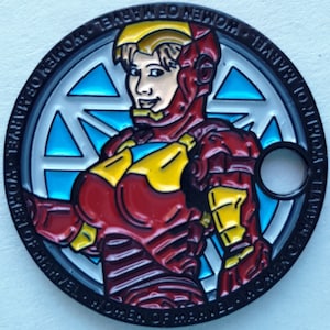 May include: A black metal pin with a red, yellow, and blue image of a female superhero wearing a helmet and armor. The pin has the text "Women of Marvel" around the edge.