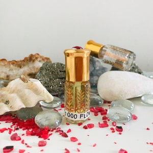 May include: A small, clear glass bottle with a gold cap and a red gemstone top. The bottle is filled with a light yellow liquid and has a label that reads "1000 FLO". The bottle is surrounded by small, red and clear glass stones.