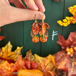 Op de afbeelding: Een paar goudkleurige oorhangers met oranje pompoenbedels en herfstkleurige bladeren. De oorbellen worden gepresenteerd tegen een groene achtergrond met herfstbladeren.
