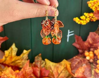 Könnte beinhalten: Ein Paar goldfarbene Ohrhänger mit orangefarbenen Kürbisanhängern und herbstfarbenen Blättern. Die Ohrringe werden vor einem grünen Hintergrund mit Herbstlaub präsentiert.
